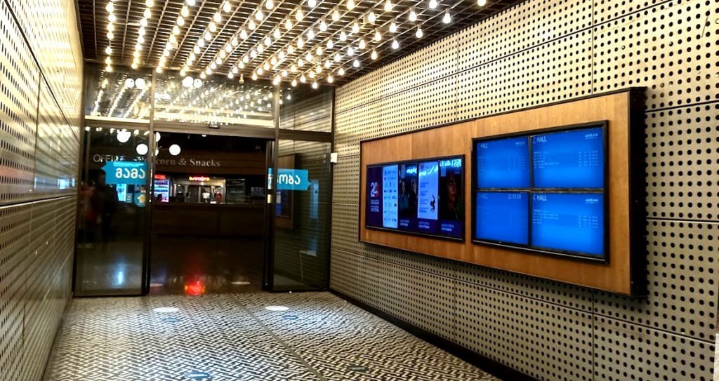 Entrance area of a modern cinema with digital display panels for the cinema programme, illuminated by a ceiling with numerous light bulbs."