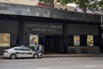 "Amirani Cinema in Tbilisi, Georgia: A modern cinema building with a police car in front of it, surrounded by trees and posters of current films."