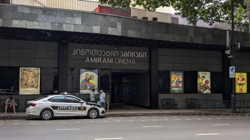 "Amirani Cinema in Tbilisi, Georgia: A modern cinema building with a police car in front of it, surrounded by trees and posters of current films."