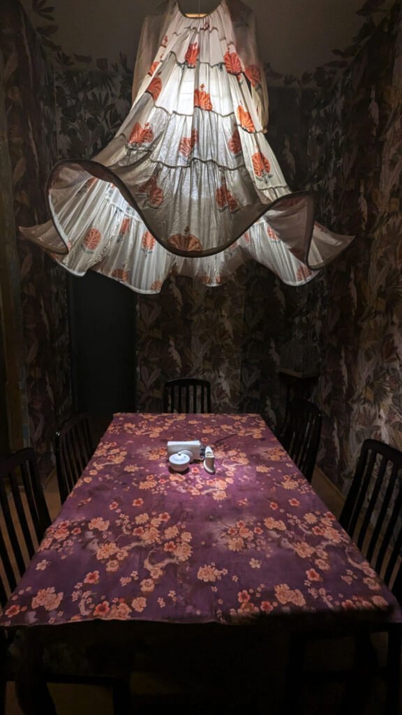 A dining table in the Nair Nairi restaurant in Tbilisi, Georgia, illuminated by a unique lampshade made from a traditional Georgian dress. The walls are covered with dark floral wallpaper.