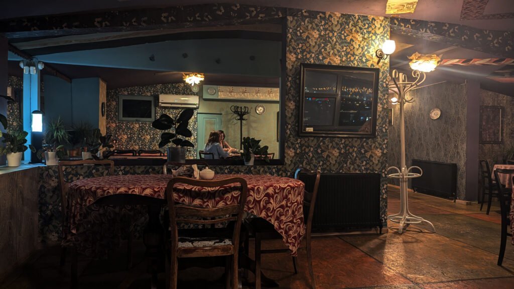 Cosy interior of the Nair Nairi restaurant in Tbilisi, Georgia. Dark wood, floral wallpaper and subdued lighting create an intimate atmosphere. A mirror reflects the dining room while guests sit at the tables and savour traditional Georgian dishes.