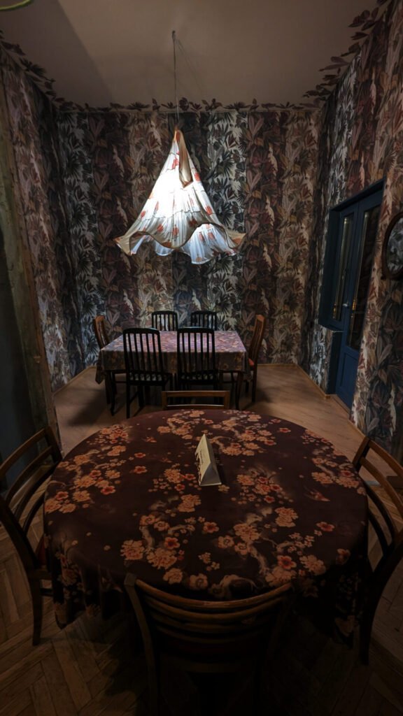 A dining area in the Nair Nairi restaurant in Tbilisi, Georgia, with a round table in the foreground and a rectangular table in the background. Both tables are covered with floral patterned tablecloths. The walls are covered with dark floral wallpaper and a chandelier made from a traditional Georgian dress hangs from the ceiling.